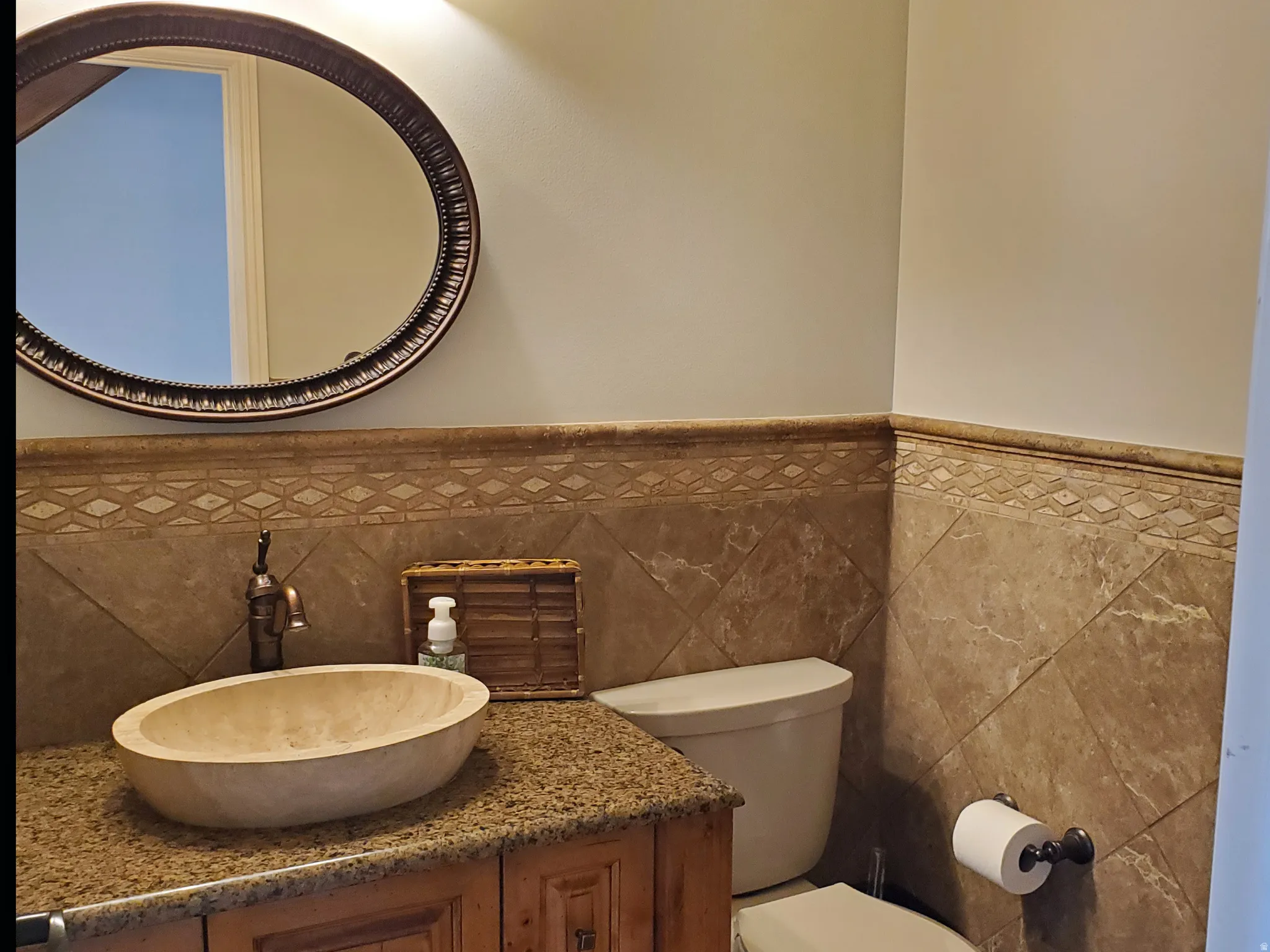 Bathroom featuring a stone vessel sink, granite countertop, wood vanity cabinetry, decorative tile wainscoting, and an oval mirror with an ornate frame