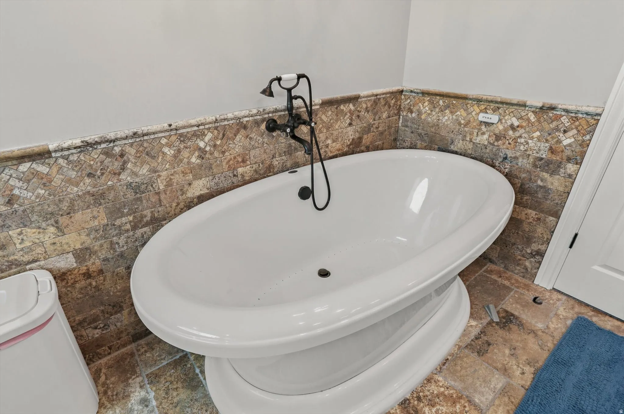 Freestanding soaking tub with a dark bronze tub filler, set against a mosaic tile wainscoting and stone-finish floor tiles