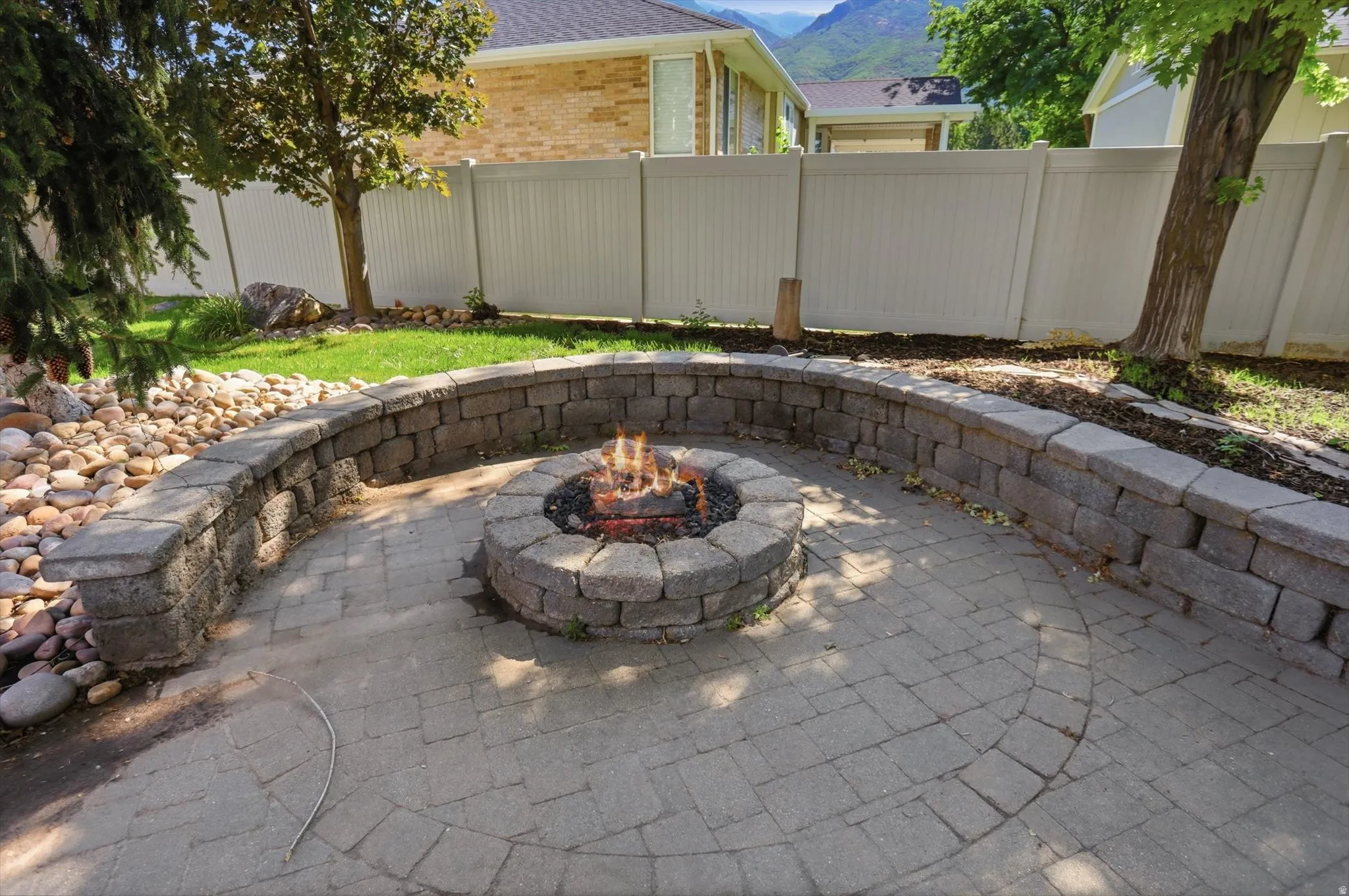 Paver patio featuring a built-in fire pit, surrounded by a curved stone retaining wall, and a pebble rock bed
