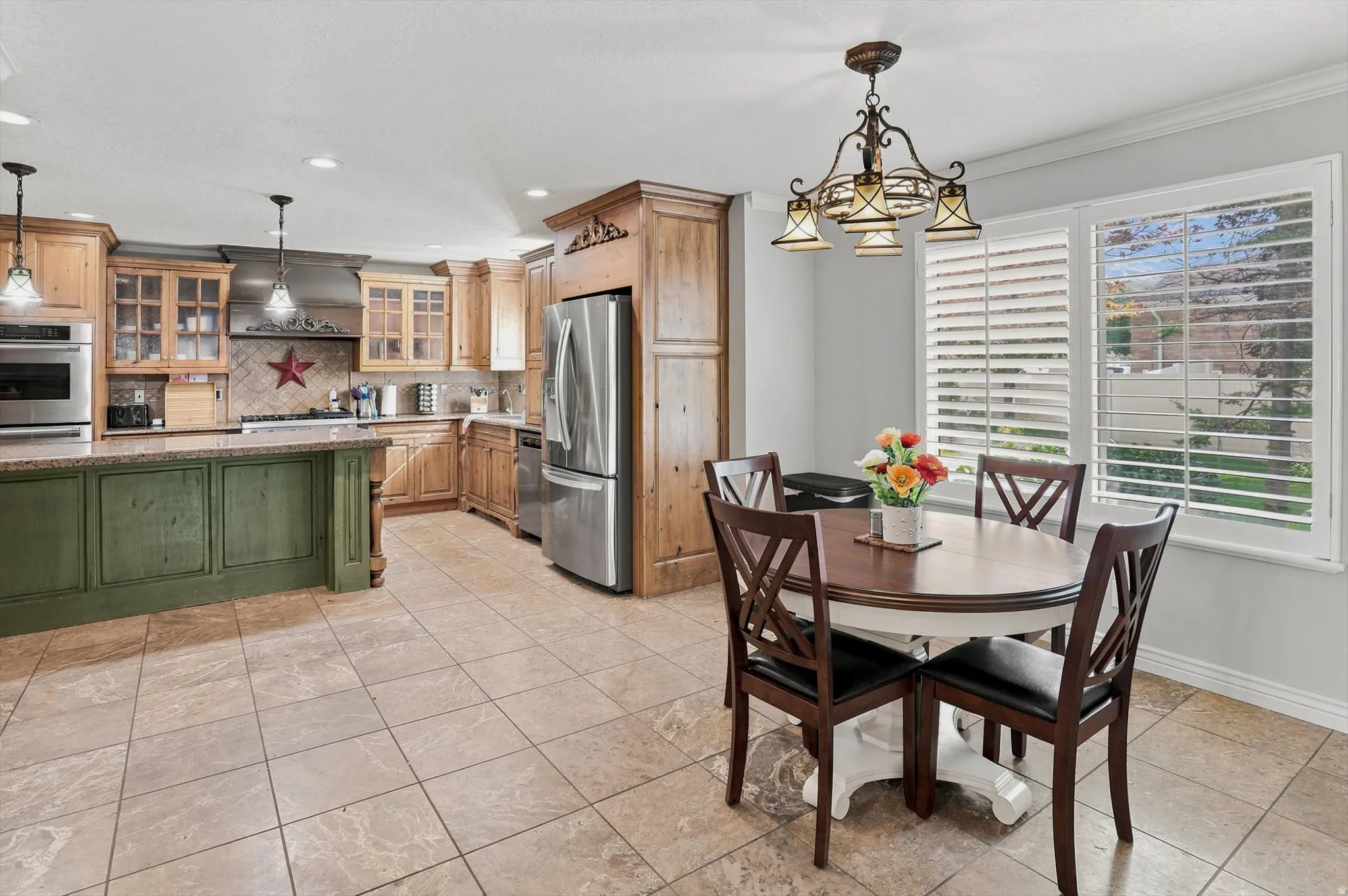 Spacious kitchen featuring tile flooring, light wood cabinetry, a green island with a stone countertop, stainless steel appliances, and a window with plantation shutters