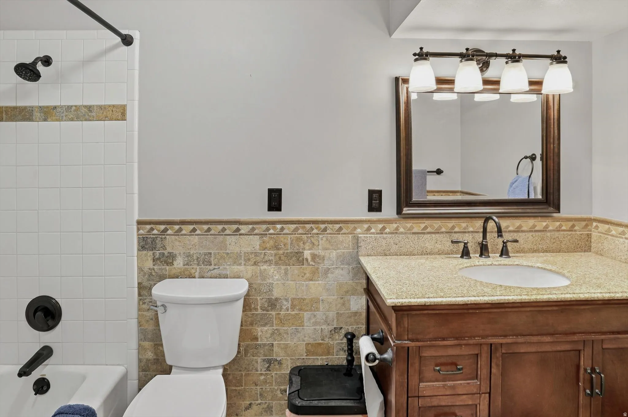Bathroom featuring a white subway tile shower with a decorative accent band, a built-in bathtub with matte black fixtures, a stone mosaic wainscoting, a vanity with a light-colored countertop and undermount sink, and a framed mirror with a four-light fixt