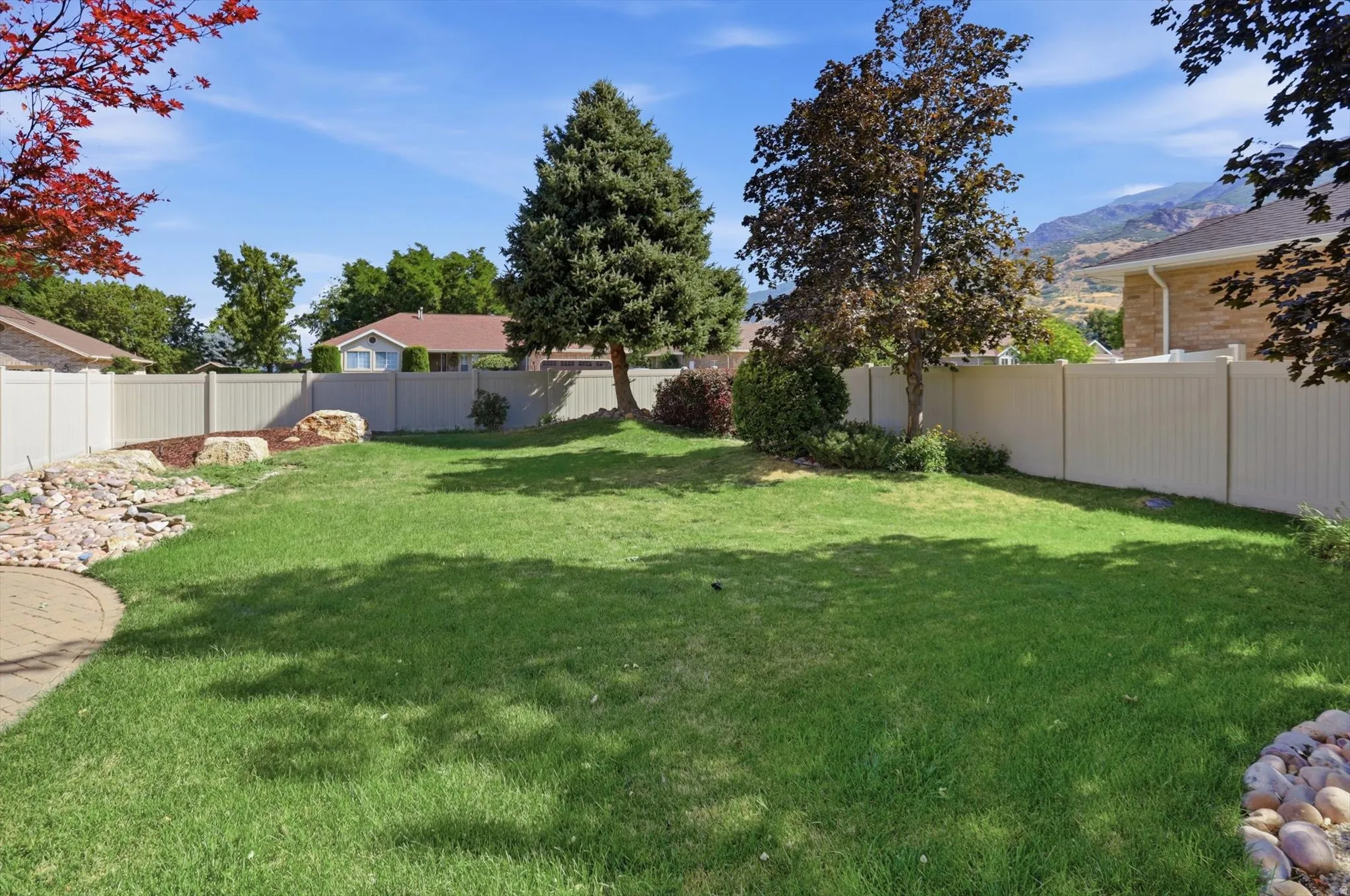 Expansive lawn area enclosed by perimeter fencing, featuring mature trees, a decorative rock garden, and a curved paver patio