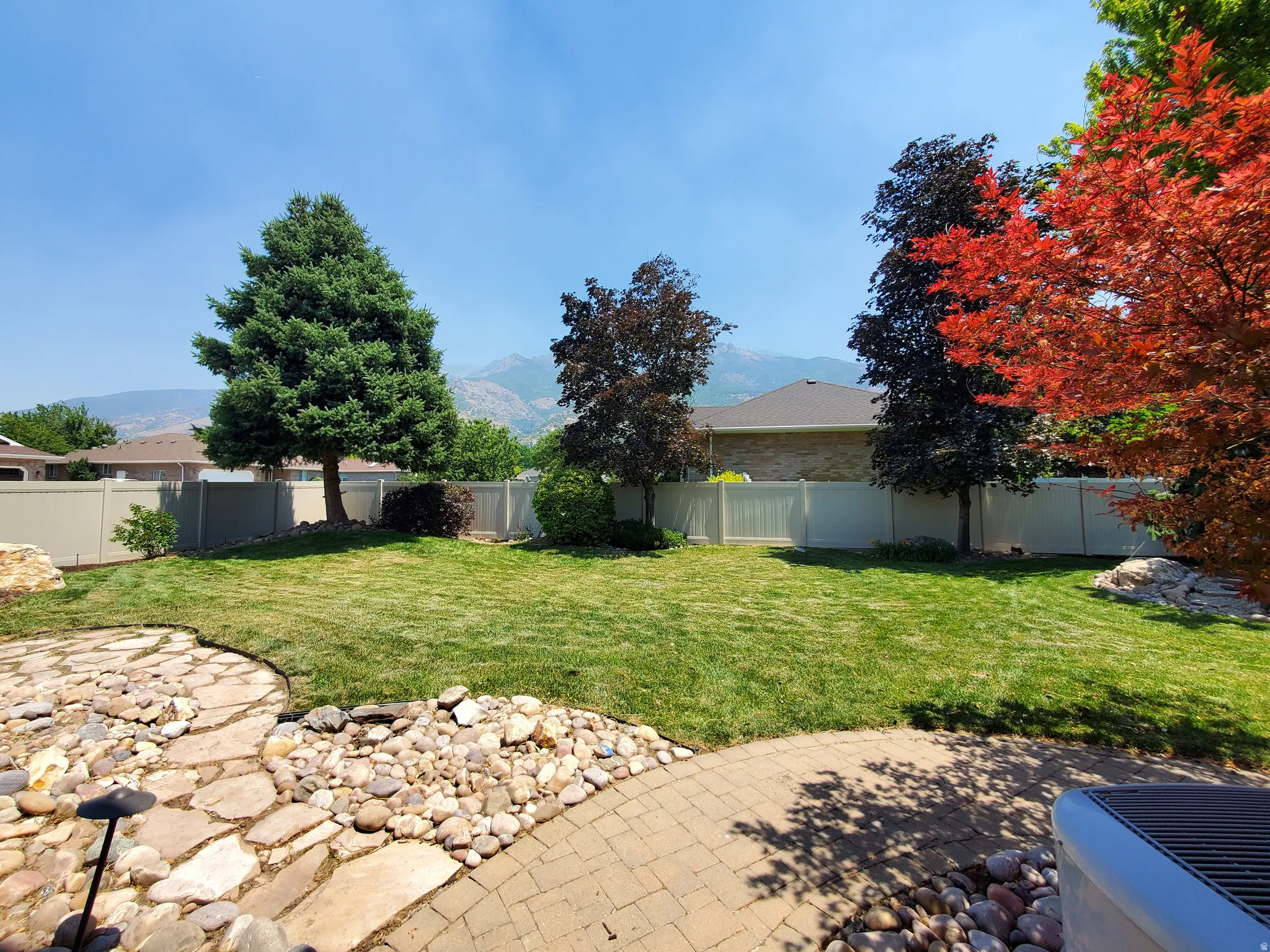Expansive backyard featuring a manicured lawn, established trees, a stone patio, and a privacy fence
