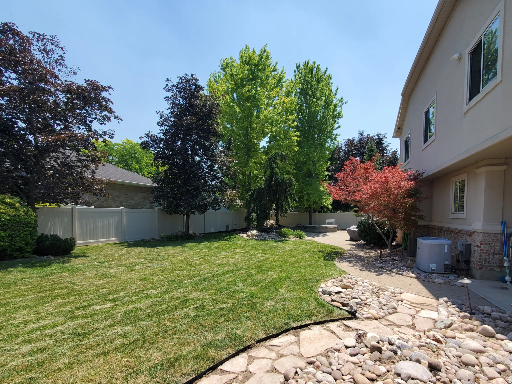 Expansive backyard featuring a manicured lawn, mature trees, a private vinyl fence, and a stone pathway leading to a paved patio area