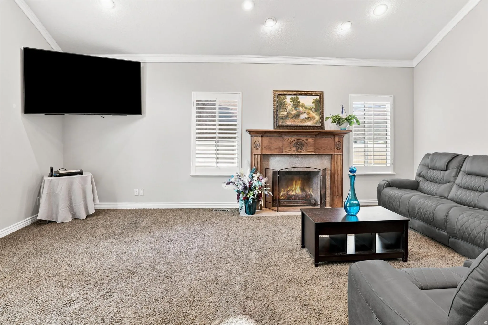 Living area featuring a wood-mantled fireplace with a metal screen, white plantation shutters, crown molding, recessed lighting, and neutral-toned carpeting