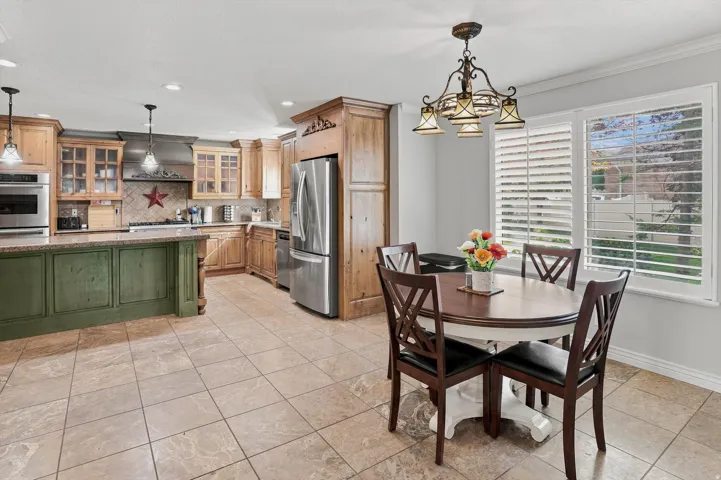 Spacious kitchen featuring tile flooring, light wood cabinetry, a green island with a stone countertop, stainless steel appliances, and a window with plantation shutters