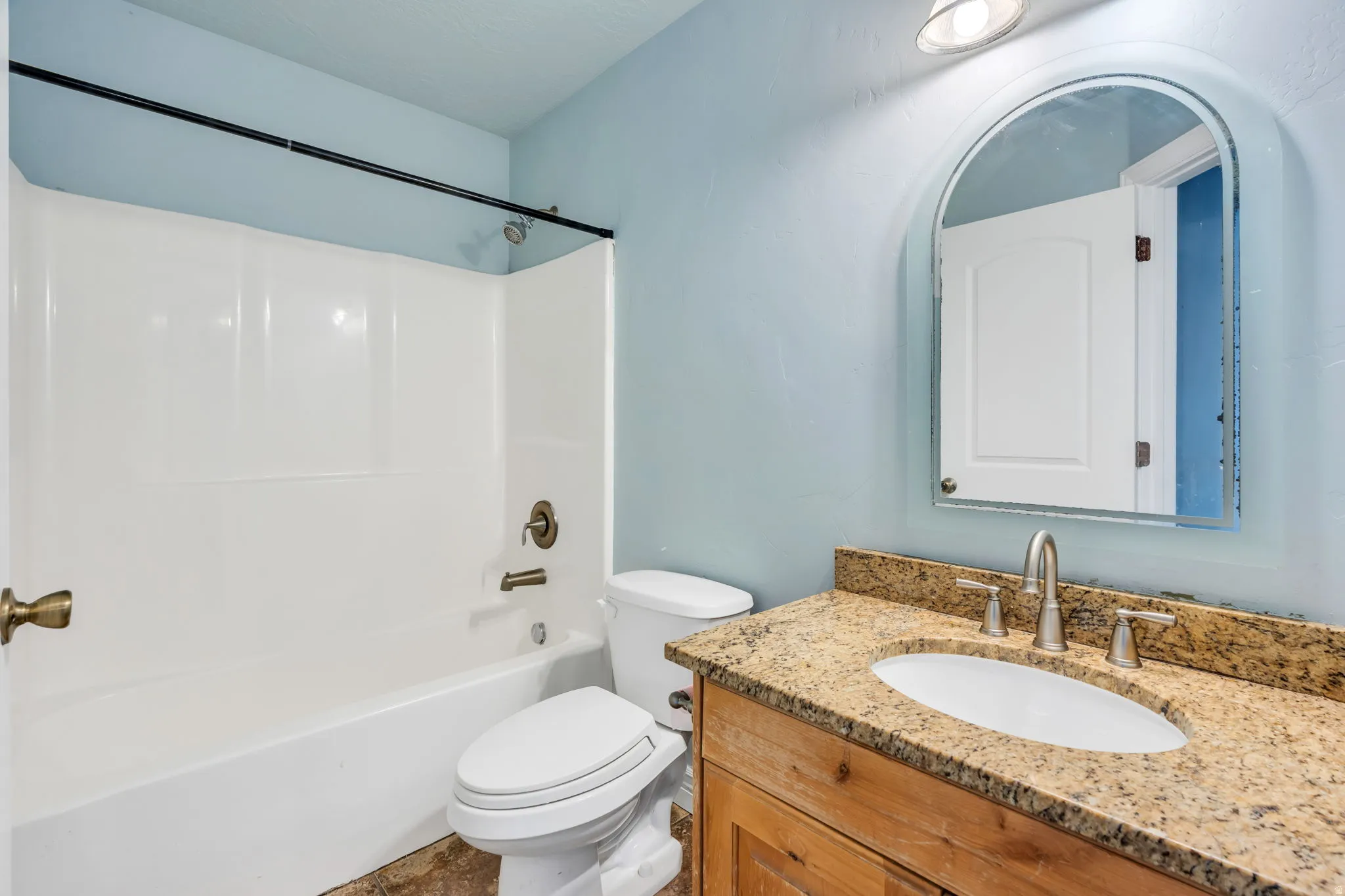 Bathroom featuring a light blue wall color, a white bathtub with a shower kit, an arched mirror, a vanity with a light wood-finish cabinet, and granite countertop with an undermount sink