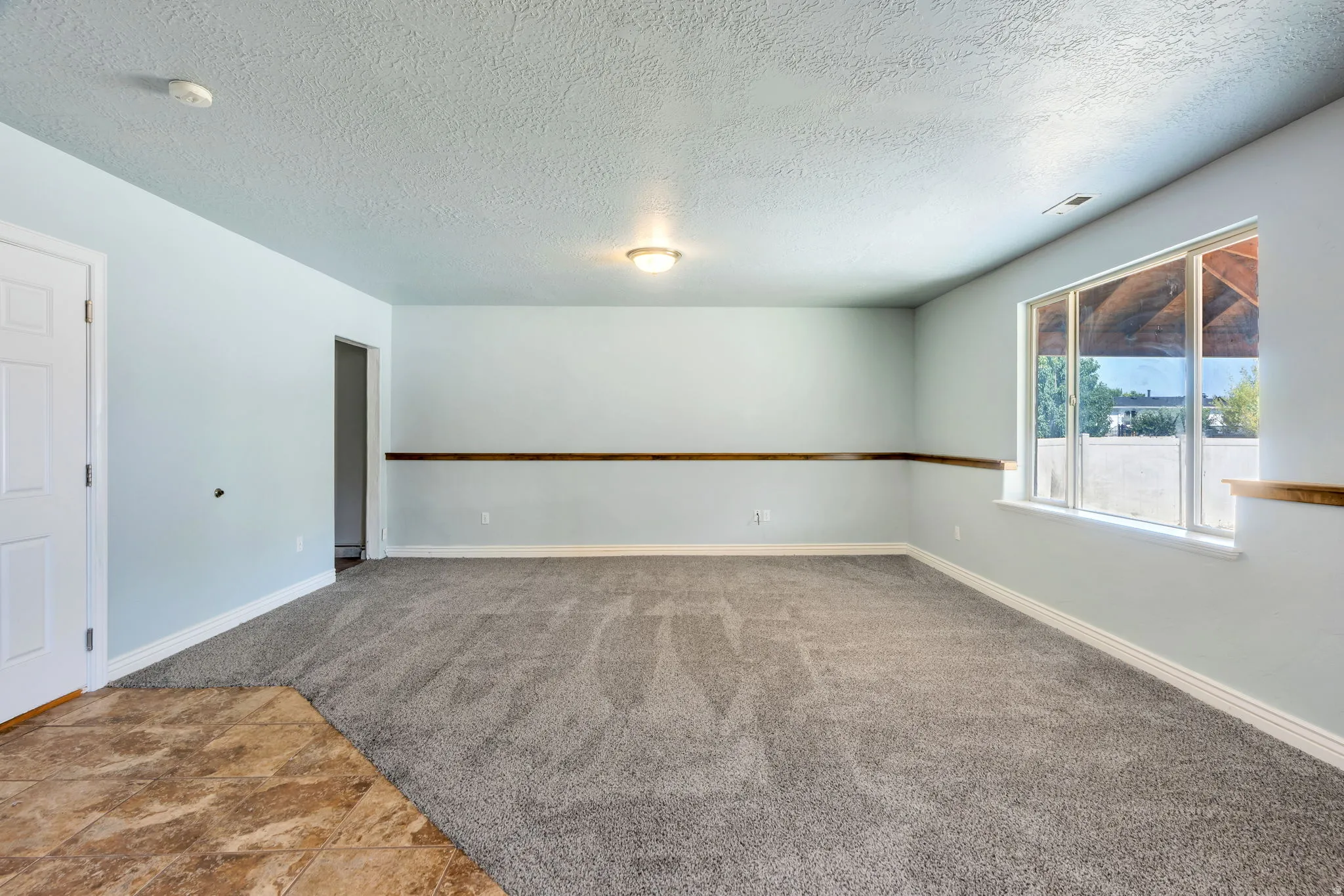 Spacious room featuring light blue walls, gray textured carpeting, and a large window with white trim