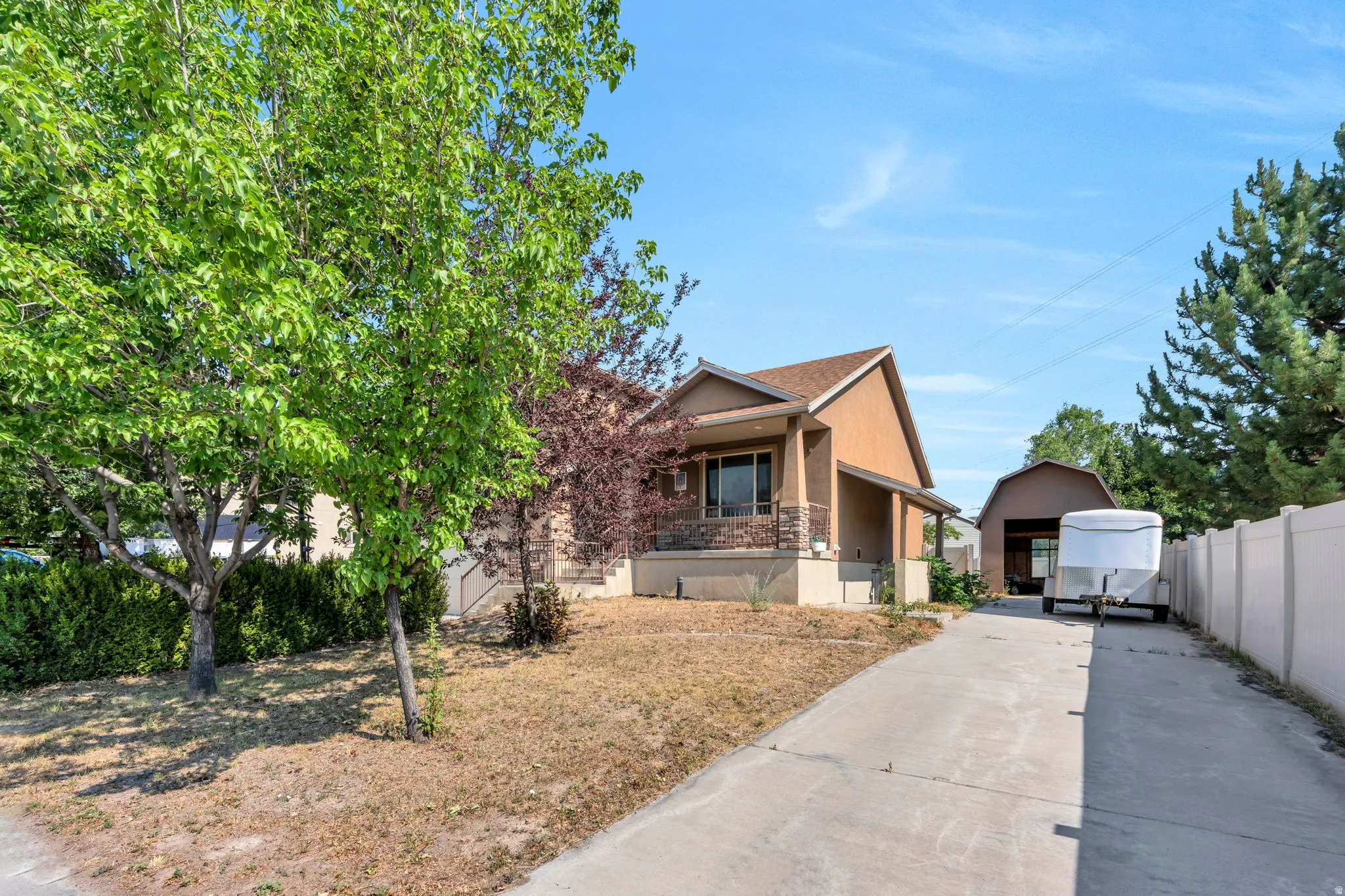 Stucco exterior residence featuring a covered front porch with stone accents, mature landscaping, and a paved driveway leading to a detached auxiliary building