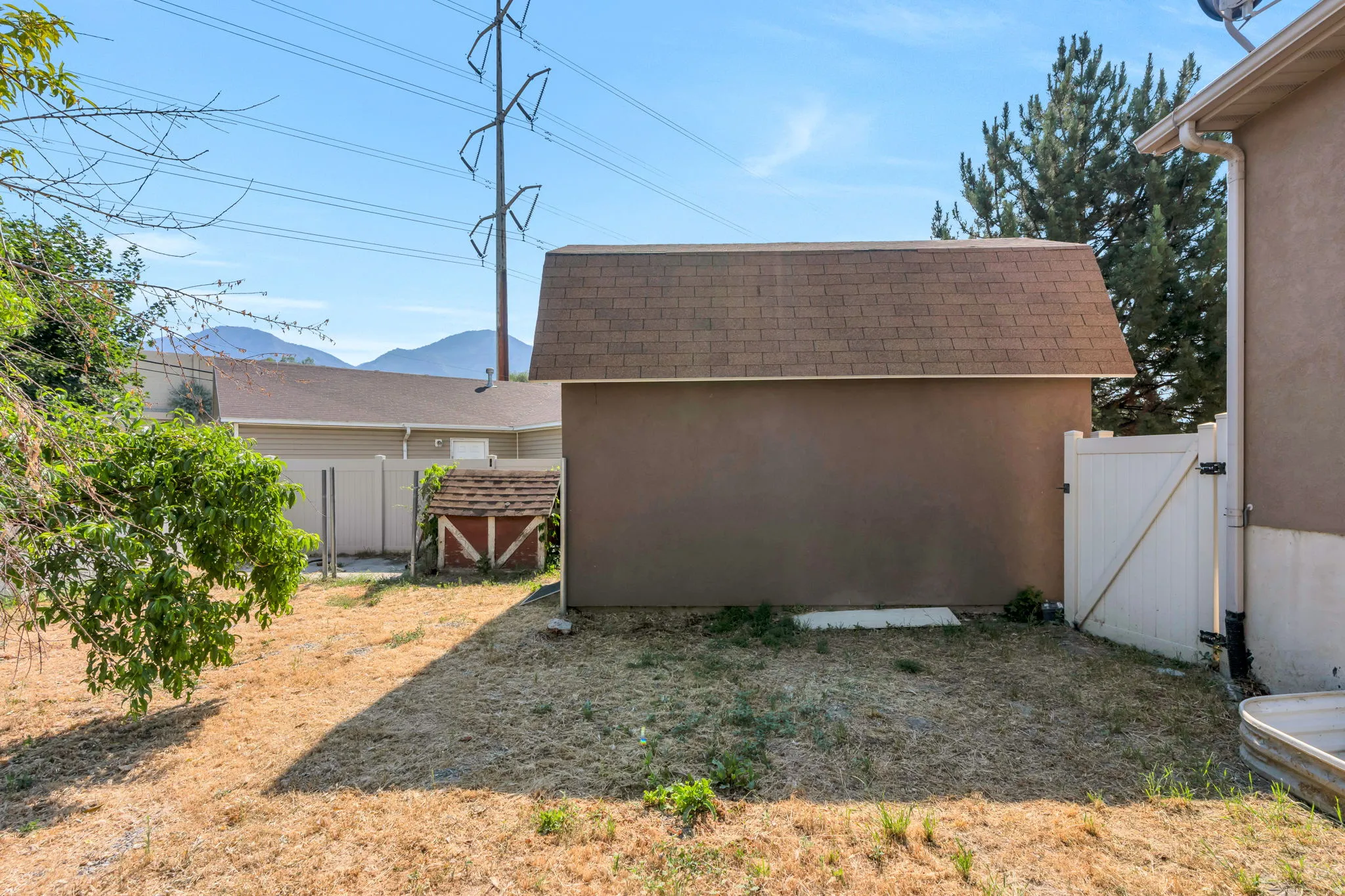 Expansive backyard featuring a brown stucco shed with a gambrel roof, a white vinyl fence, and a white vinyl gate