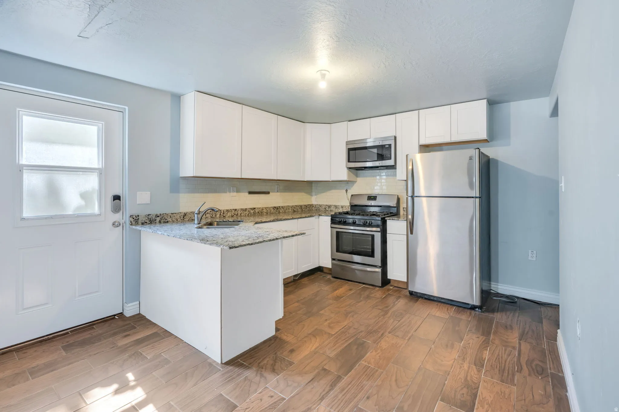 Full kitchen with white cabinetry, stainless steel appliances, granite  countertops, wood-finish flooring, and a white paneled door with frosted glass inserts