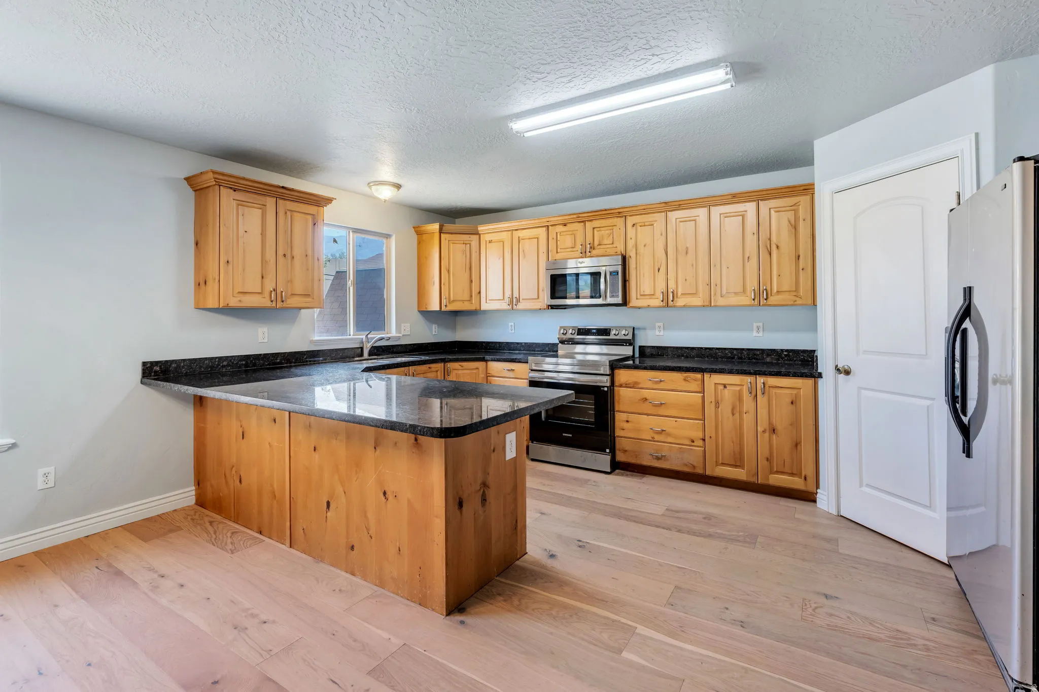 Kitchen featuring wood cabinetry with black granite countertops, a central island with countertop seating, stainless steel appliances, and light wood-finish flooring