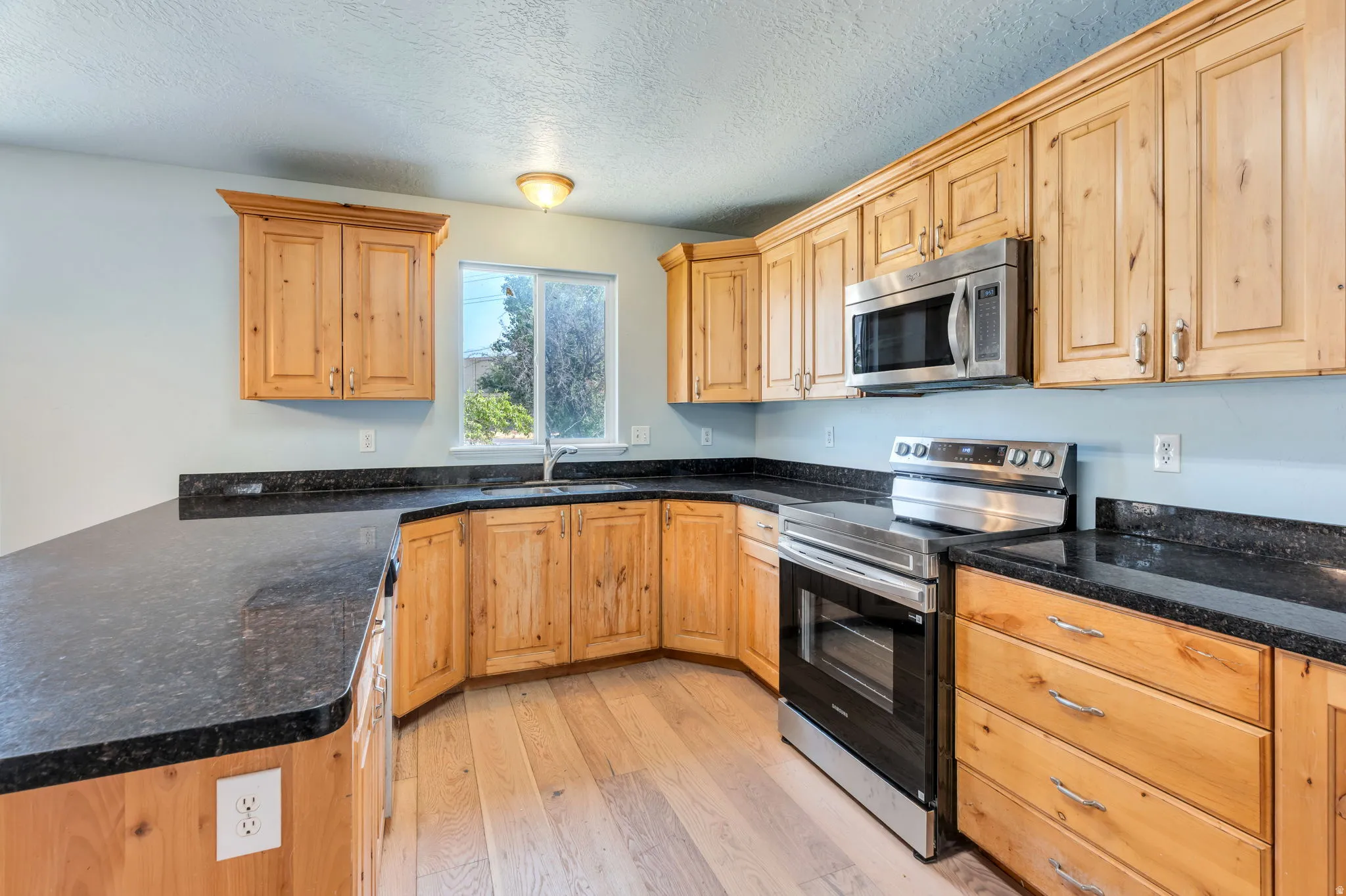 Kitchen featuring light wood cabinetry, dark granite countertops, stainless steel appliances, and wood-finish flooring