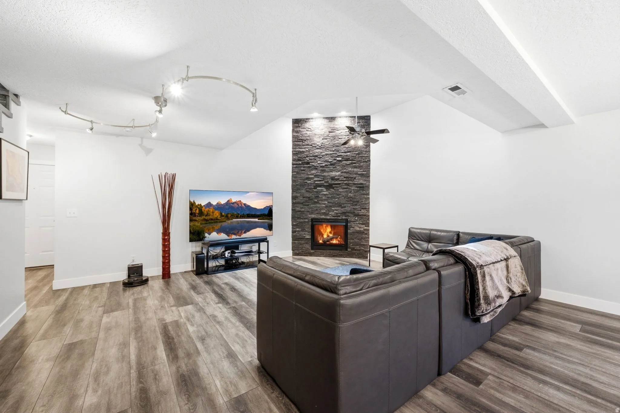 Living area featuring a stone accent wall with a built-in fireplace, vaulted ceilings, wood-finish flooring, a ceiling fan, and recessed track lighting