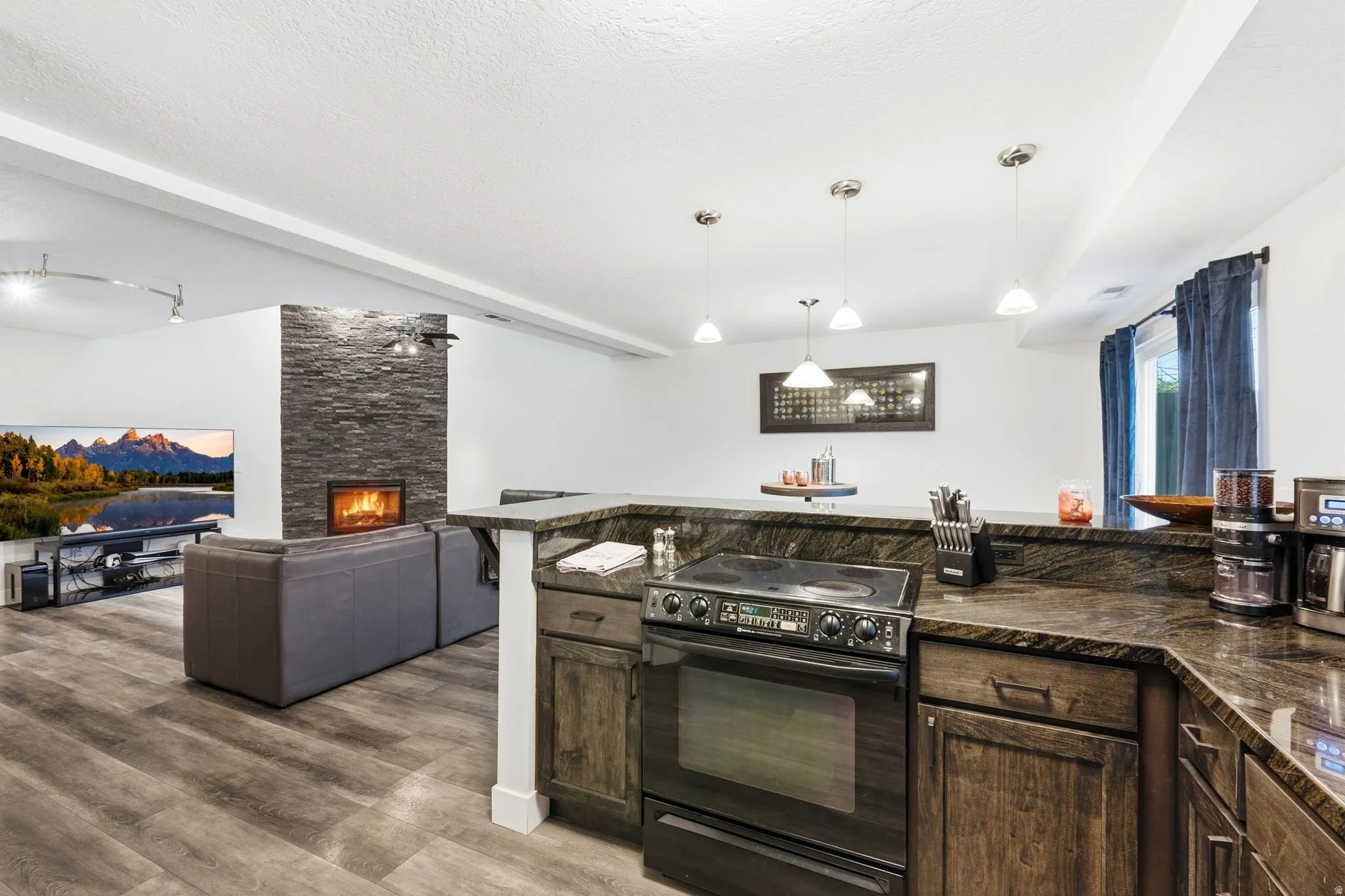 Open-concept lower level featuring a stacked stone fireplace, wood-finish flooring, and a kitchen island with stone countertops and integrated cooktop