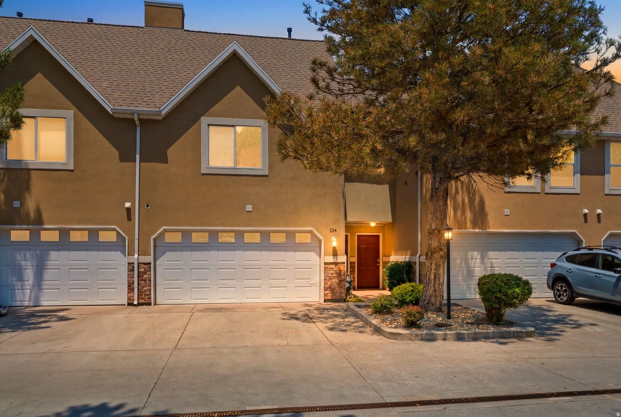 Stucco townhouse featuring a brown shingle roof, two attached garages with panel doors and upper window panels, and a prominent mature tree