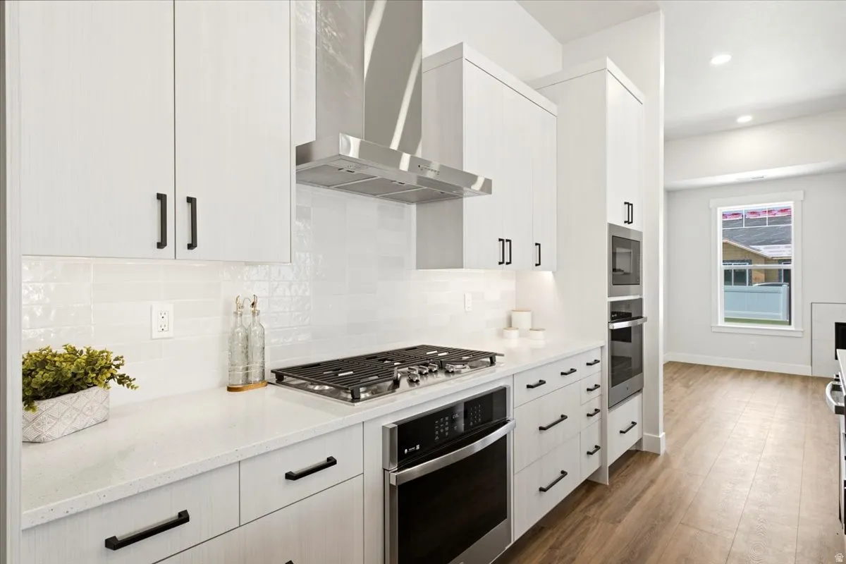 Kitchen featuring white cabinetry with matte black hardware, a stainless steel range hood, a built-in oven and microwave, white countertops, and wood-finish flooring