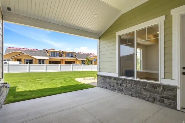 Covered concrete patio featuring a textured stone veneer foundation, light green siding, and a white vinyl fence enclosing a manicured lawn