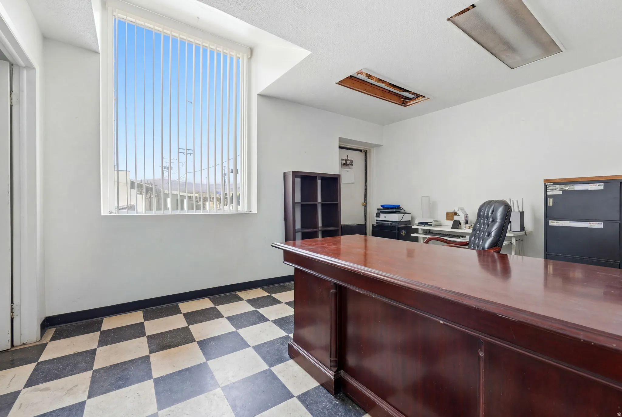 Spacious room featuring a large window with vertical blinds, checkerboard floor tiling, and a built-in wood-finish desk