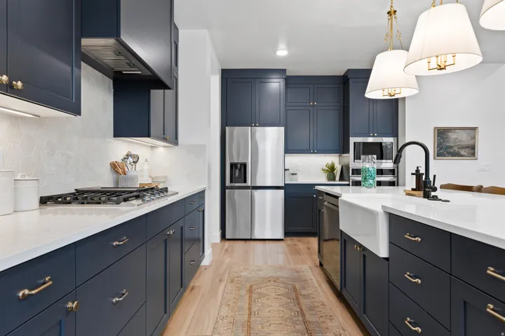 Modern kitchen featuring navy cabinetry with brass hardware, a white farmhouse sink, stainless steel appliances, light wood-finish flooring, and pendant lighting fixtures