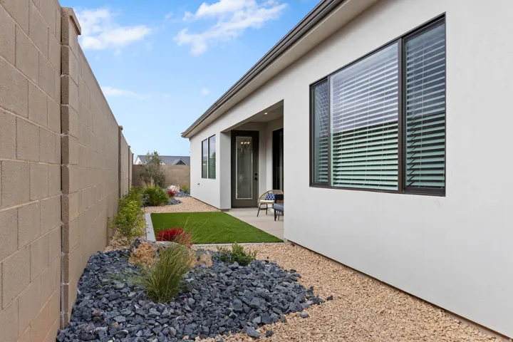 Modern stucco exterior featuring large windows with blinds, a covered patio, artificial turf, and low-maintenance xeriscaping with decorative rocks