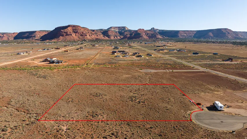 Expansive desert lot featuring a red rock mountain backdrop, scattered vegetation, and distant residential structures