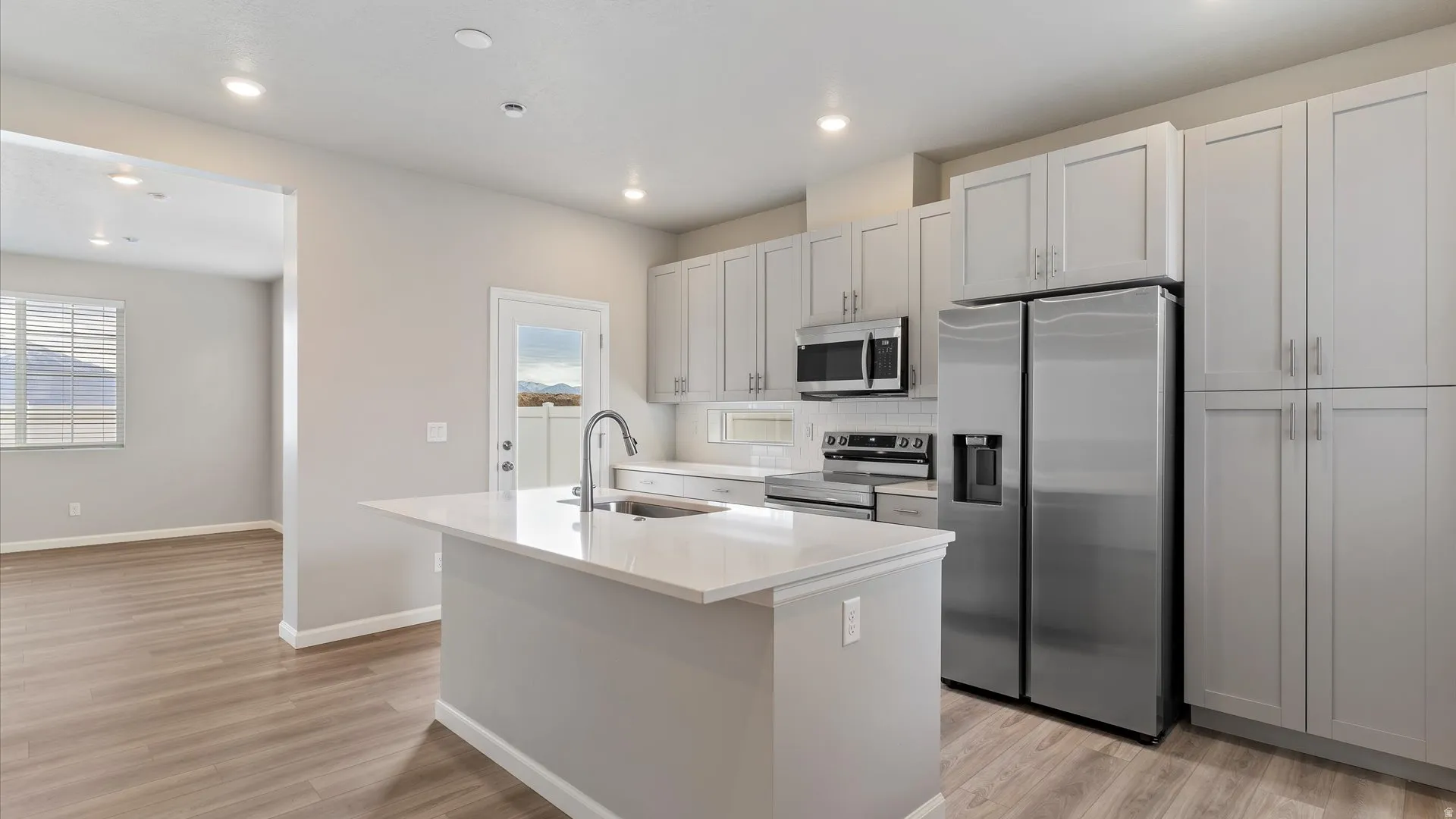 Spacious kitchen featuring a central island with an undermount sink, stainless steel appliances, light gray shaker cabinetry, wood-finish flooring, and recessed lighting