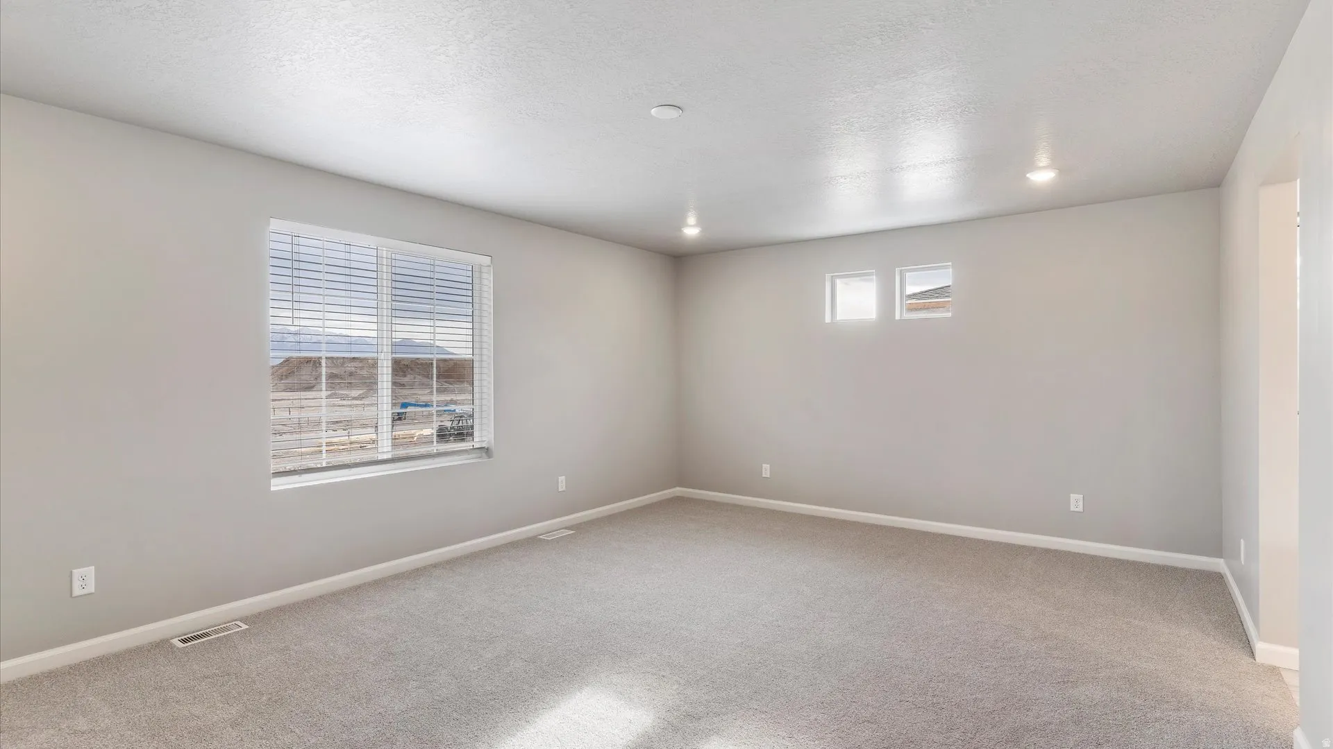 Expansive room featuring light gray carpeting, recessed lighting, a large window with blinds, two smaller square windows, and neutral wall paint