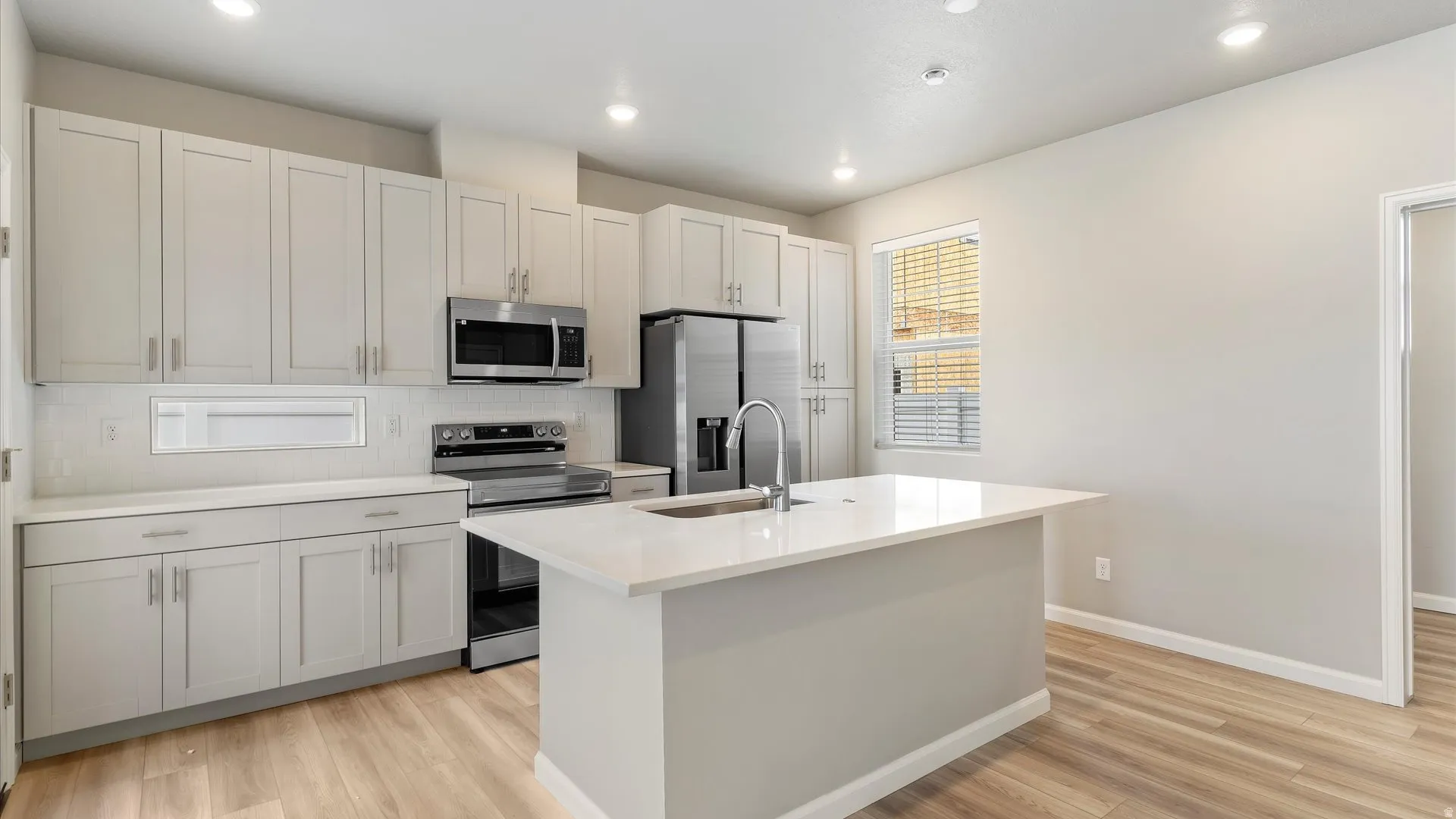 Modern kitchen featuring light gray cabinetry, stainless steel appliances, a white subway tile backsplash, a central island with a sink, and wood-finish flooring
