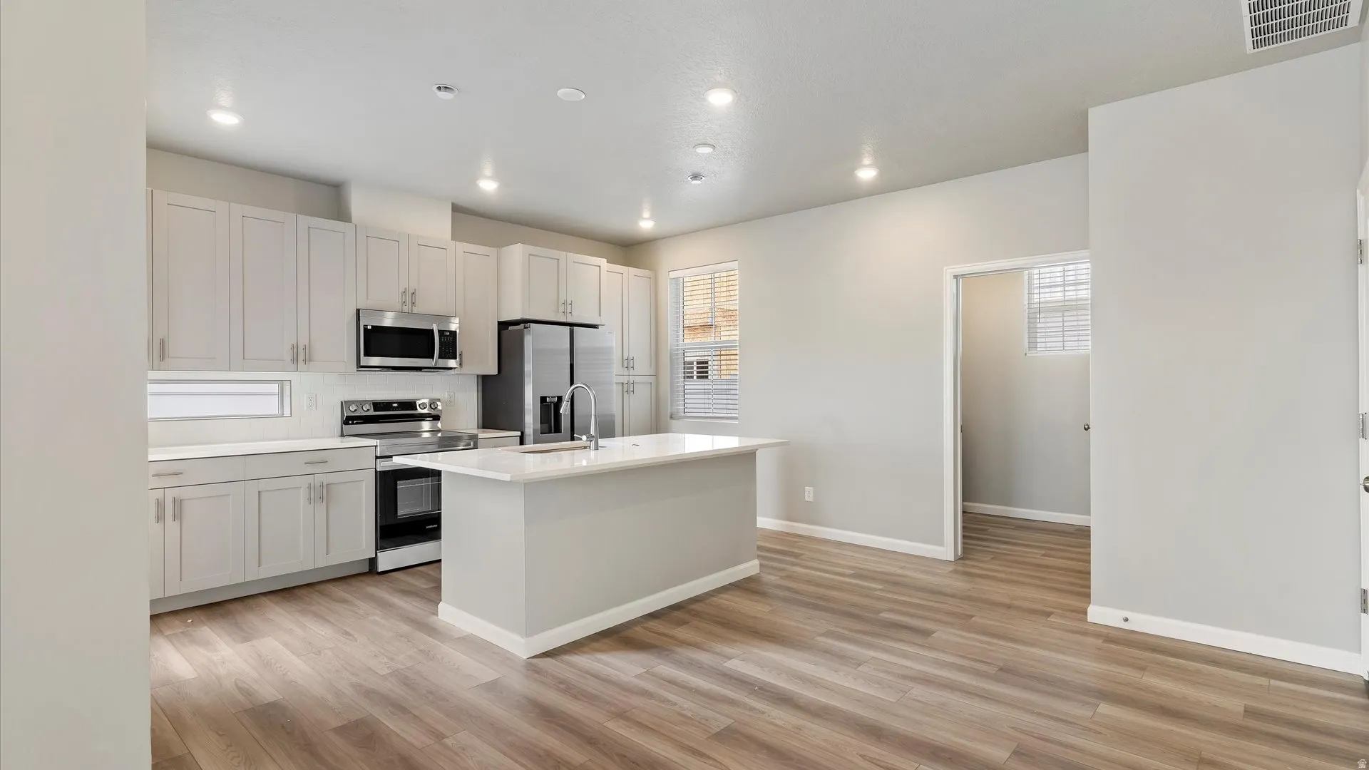 Modern kitchen featuring light gray cabinetry, stainless steel appliances, a center island with sink, wood-finish flooring, and recessed lighting
