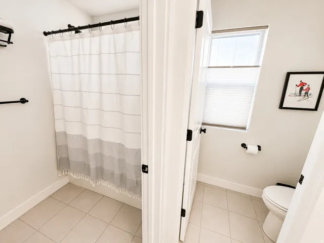 Bathroom featuring light-toned tile flooring, a shower area with a black shower rod, and a separate toilet area with a window