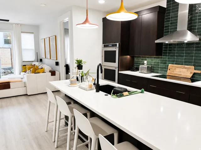 Modern kitchen featuring a large white island with seating, a black undermount sink, dark wood cabinetry, a green subway tile backsplash, and stainless steel double ovens