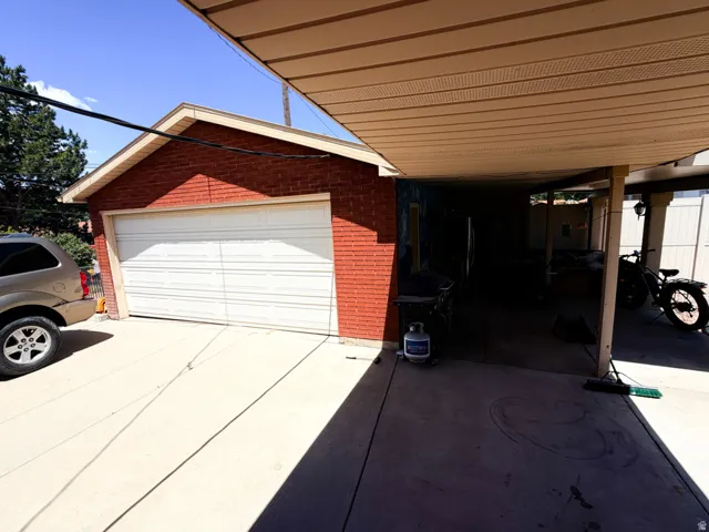 Red brick garage structure with a white panel garage door, concrete driveway, and an attached carport featuring a beige metal ceiling