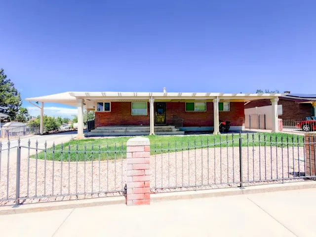 Brick facade residence featuring a covered carport, front yard with gravel and grass, wrought iron fence with brick pillars, and a visible front entry