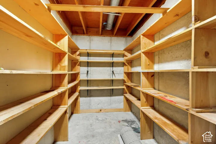 Utility space featuring extensive wood shelving, concrete walls, concrete flooring, and exposed ceiling joists