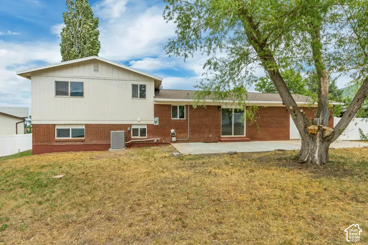 Rear exterior featuring brick and siding finishes, multiple windows, a ground-level patio, and a mature tree