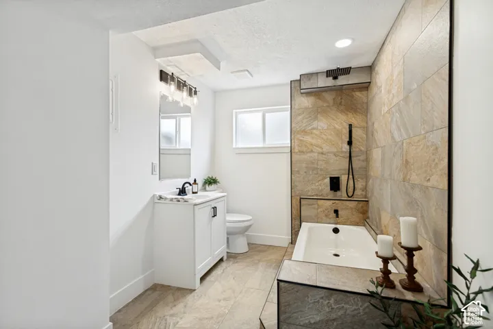 Bathroom featuring a soaking tub with integrated tiled surround, large format stone-finish wall tiling, a rainfall shower fixture, and a white vanity with a dark countertop and black hardware