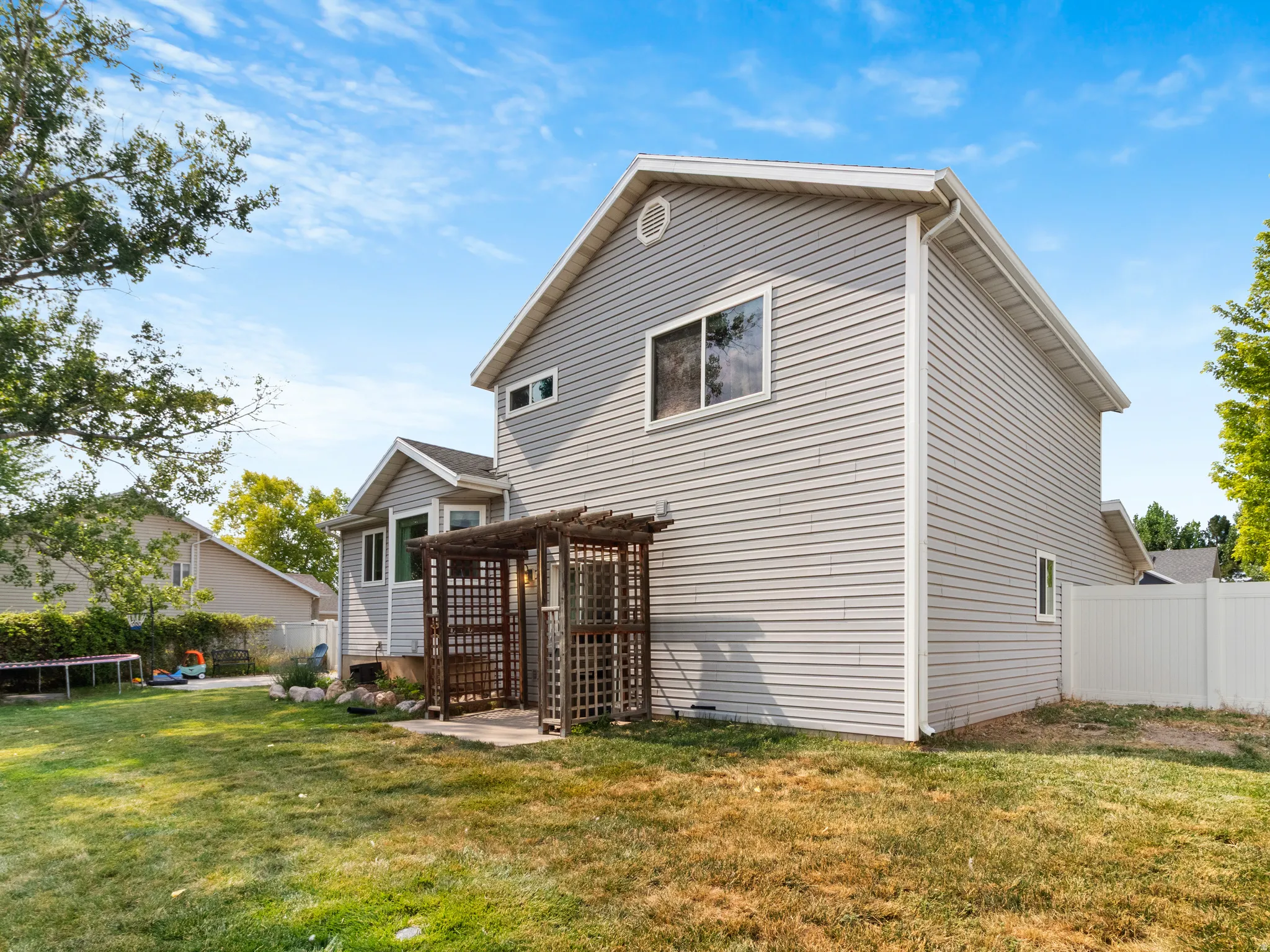 Rear exterior showcasing light gray siding, multiple windows, a ground-level concrete patio with a wooden pergola structure, and a green lawn