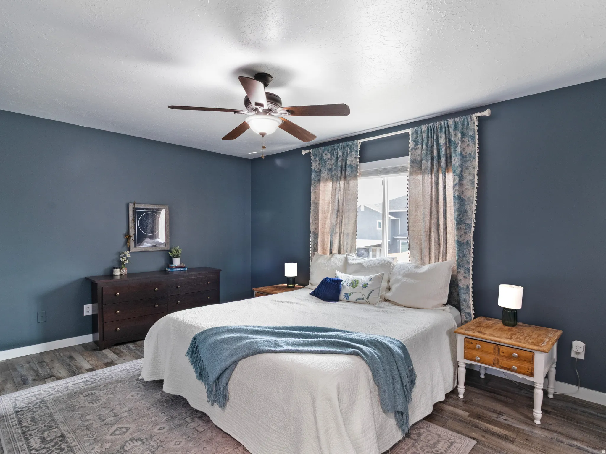 Spacious interior room featuring wood-finish flooring, a window with decorative valances and curtains, a ceiling fan with light fixture, and dark blue wall paint