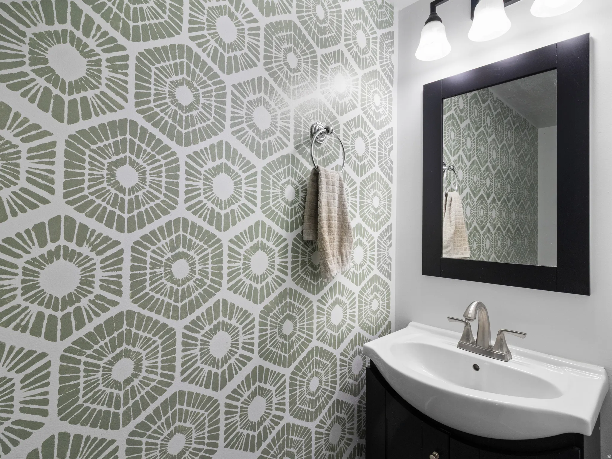 Powder room featuring a patterned accent wall, a vanity with an integrated sink, a framed mirror, a wall-mounted light fixture, and a towel ring