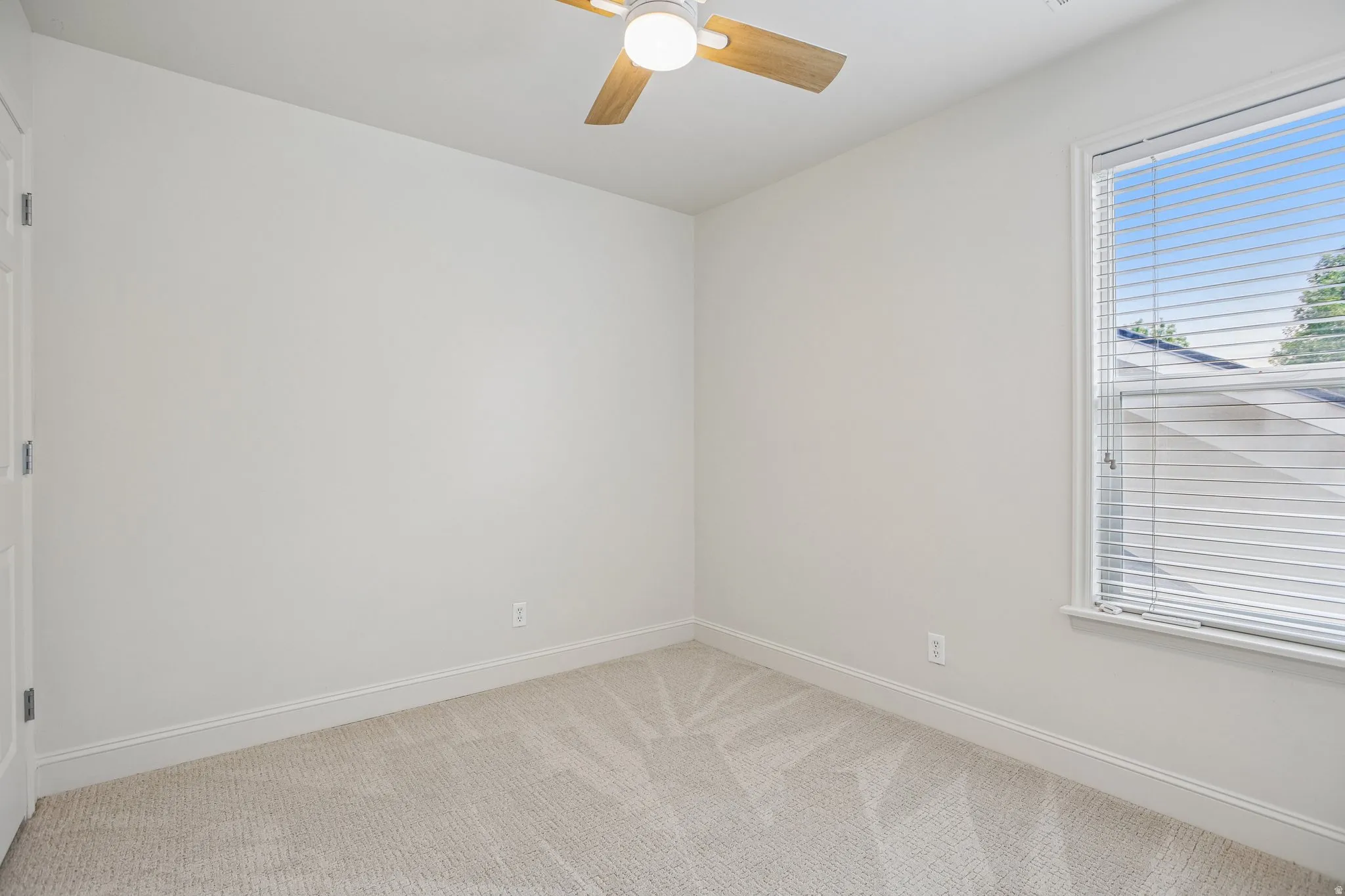Bright room featuring plush neutral-toned carpeting, a ceiling fan with wood-finish blades, and a window with blinds