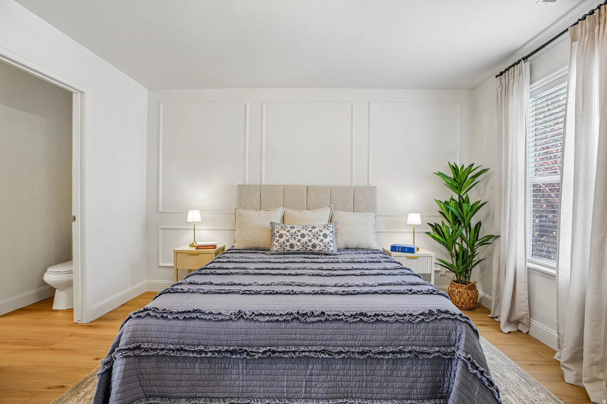 Bedroom featuring wood-finish flooring, a decorative accent wall with panel molding, and a window with blinds and drapes