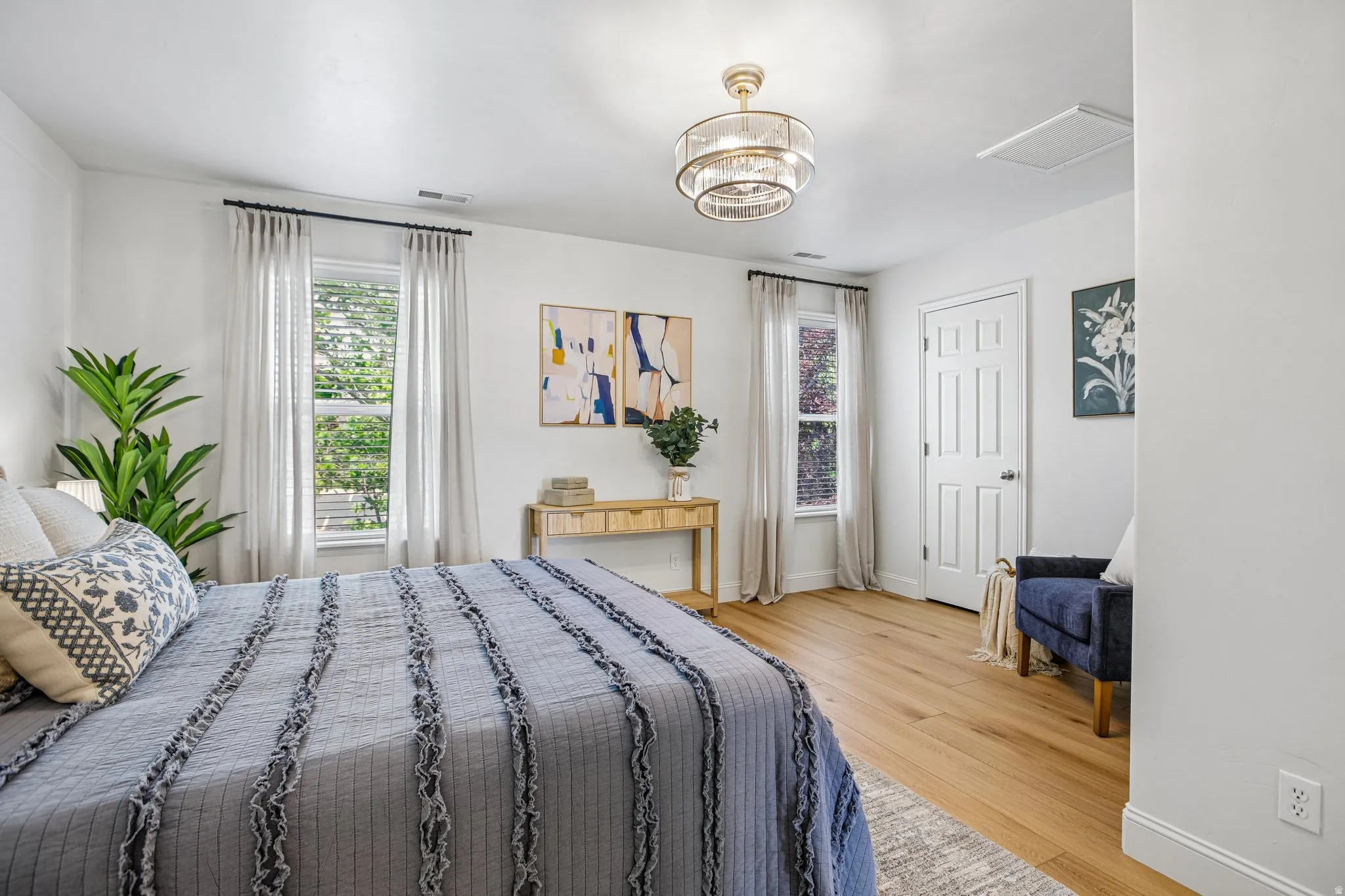 Spacious room featuring wood-finish flooring, two windows with blinds, a multi-tiered chandelier, and a paneled door