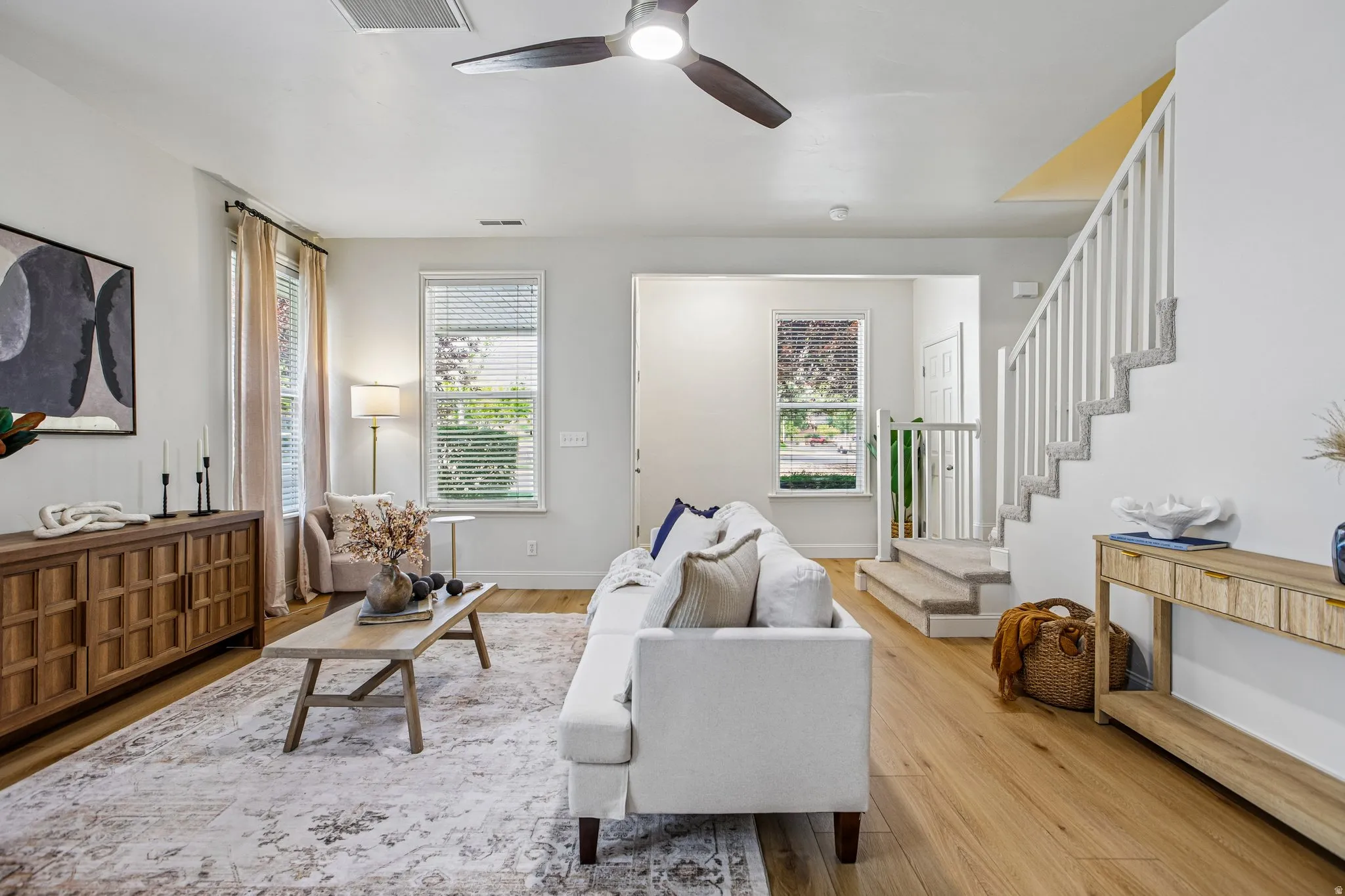 Spacious living area featuring wood-finish flooring, a contemporary ceiling fan, multiple windows with blinds, and a carpeted staircase with white railings
