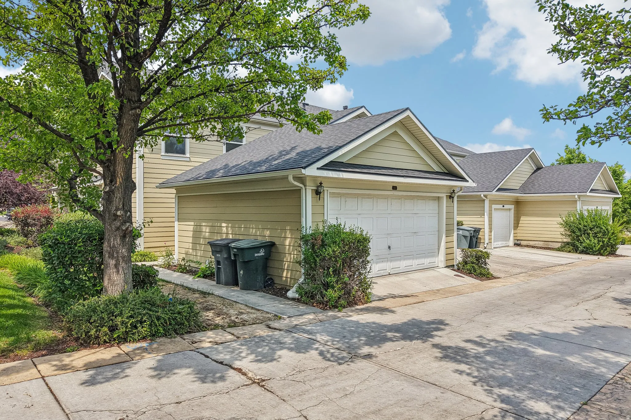 Exterior featuring light-colored siding, a grey shingled roof, an attached garage with a white door, established trees, and concrete paving