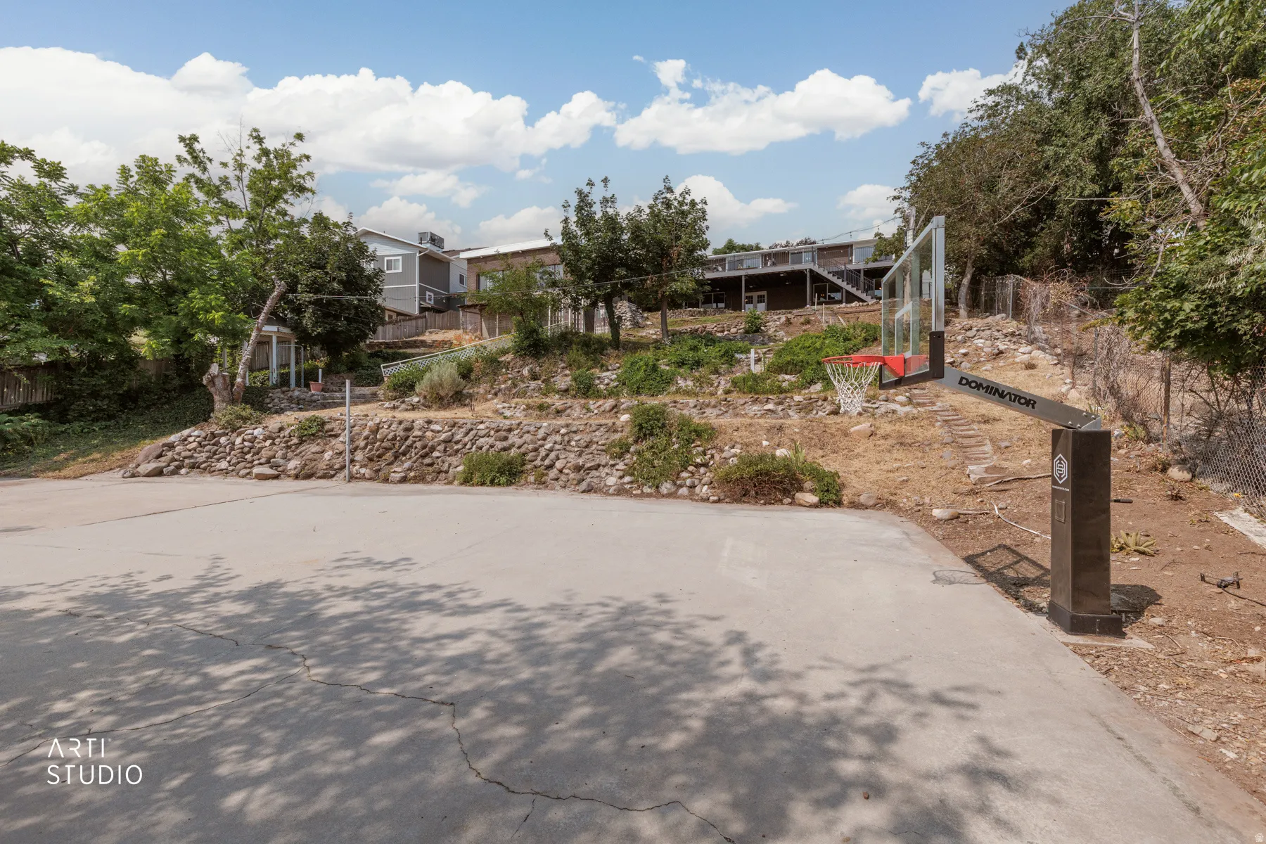 Paved basketball court featuring basketball hoops with a clear backboard and red rim, surrounded by natural landscaping and mature trees