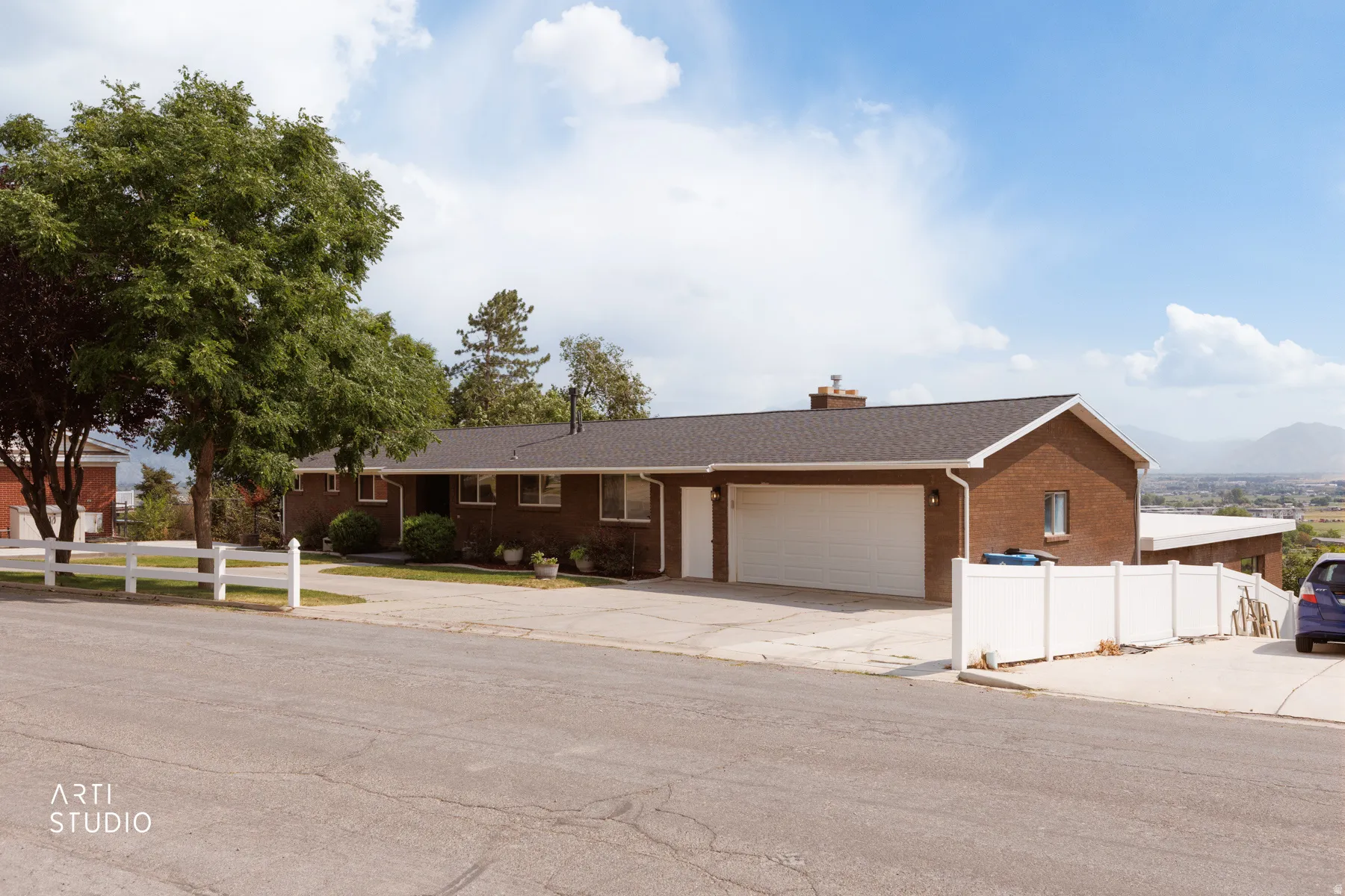 Brick exterior residence featuring a two-car garage, white vinyl fencing, a mature tree, and a paved driveway