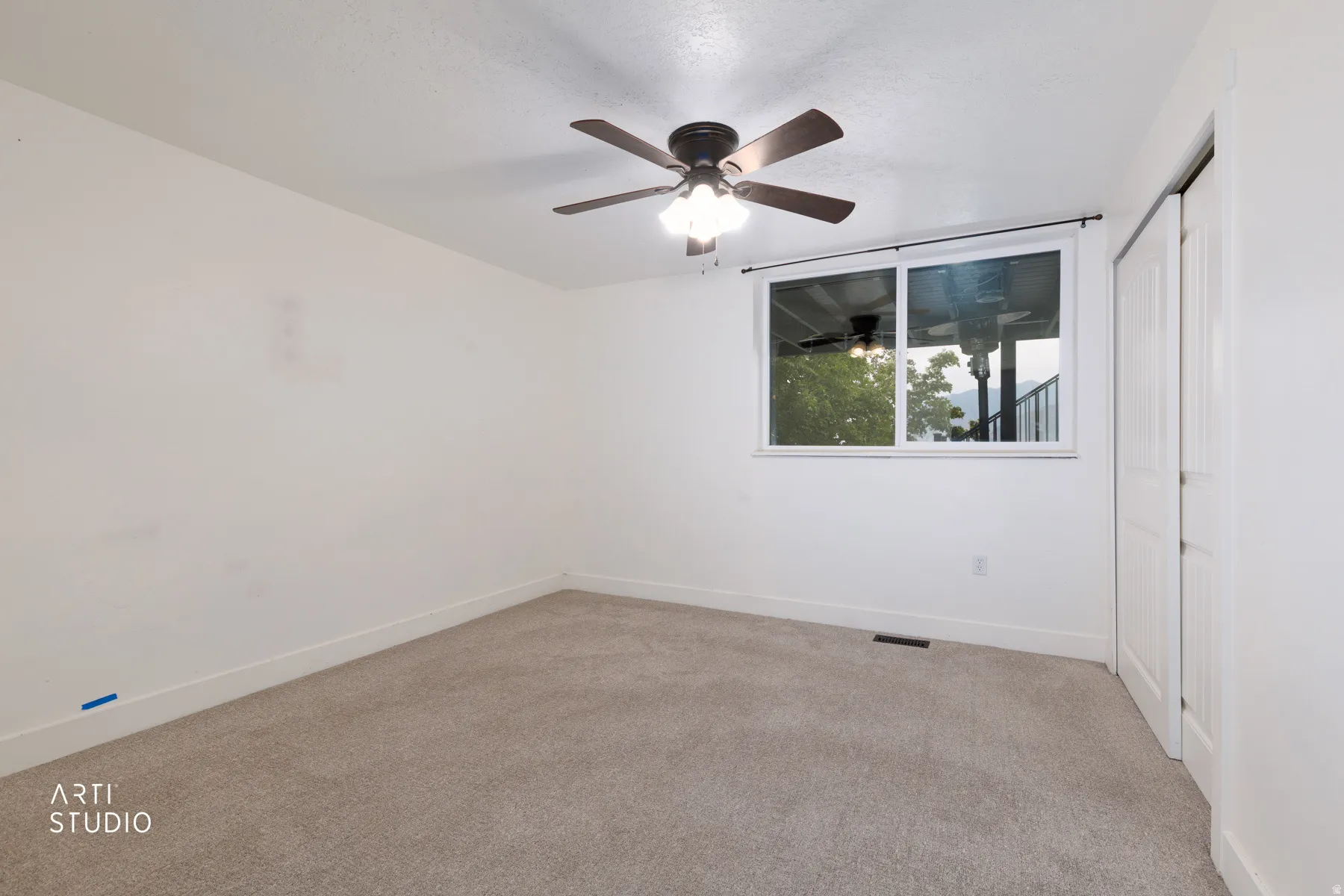 Carpeted interior room featuring a window overlooking an exterior balcony, a ceiling fan with integrated lighting, and white bi-fold closet doors
