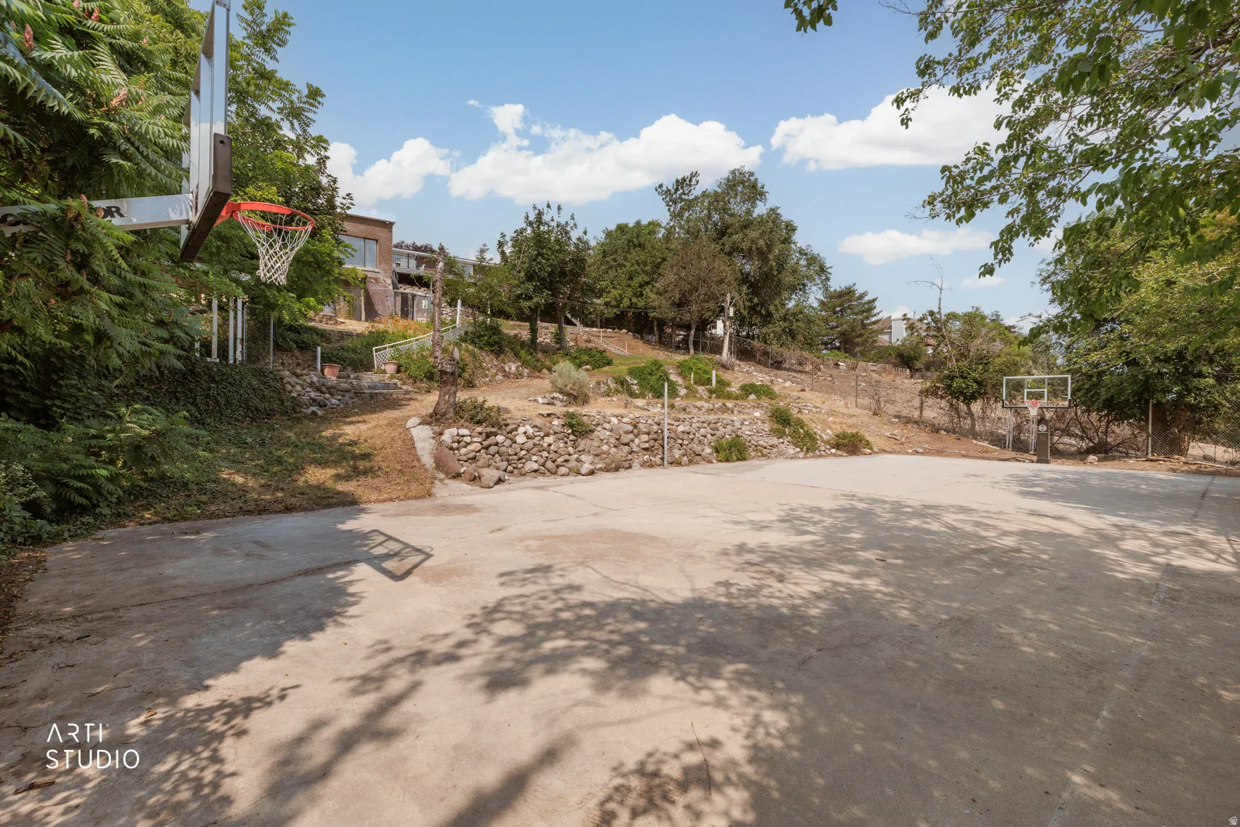 Paved outdoor court with two basketball hoops, surrounded by mature trees and natural landscaping