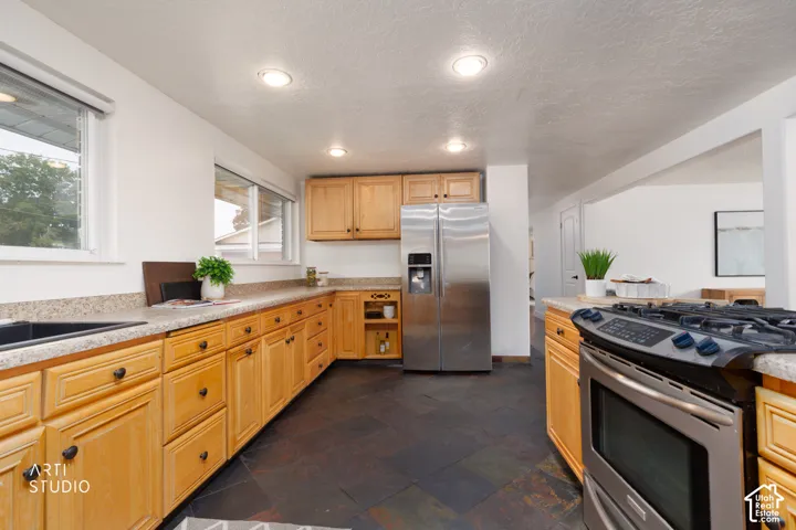 Kitchen featuring natural wood cabinetry, dark stone-finish flooring, a stainless steel side-by-side refrigerator, and a stainless steel gas range